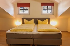 A double bed with two bedside lamps, a window with a red valance, and a sloped ceiling. - Holiday apartment with 2 bedrooms (Bodenmais)