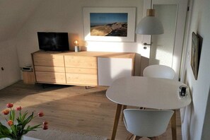 Ein Zimmer mit einem Holzkoffer, einem Fernseher, einem weißen Tisch mit Stühlen, einer Lampe und einem gerahmten Bild an der Wand.