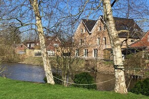 A traditional brick house with a river in the foreground, surrounded by trees and a grassy area. - Holiday apartment with a view of the sluice (Neuharlingersiel)