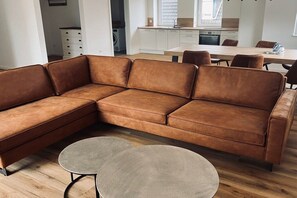 Een moderne woonkamer met een bruine leren hoekbank, een ronde salontafel en een eetgedeelte met een houten tafel en stoelen.