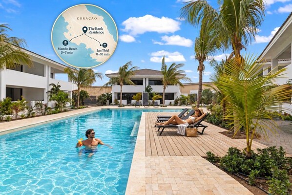 A swimming pool area with lounge chairs and palm trees, surrounded by modern buildings. - The Bank | Poolside Apartment. | Mambo & Jan Thiel (Willemstad)