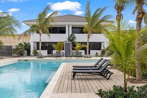 A modern two-story house with a swimming pool, lounge chairs, and palm trees. - The Bank | Poolside Apartment. | Mambo & Jan Thiel (Willemstad)