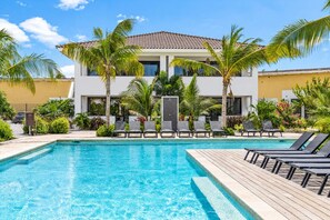 A swimming pool with lounge chairs and palm trees, surrounded by a building with a tiled roof. - The Bank | Poolside Apartment. | Mambo & Jan Thiel (Willemstad)