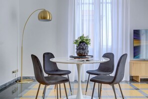 Een eethoek met een ronde tafel, vier stoelen, een vaas met bloemen, een vloerlamp en een schilderij aan de muur.