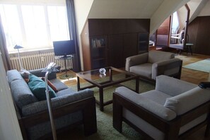 Un salon comprenant un canapé, deux fauteuils, une table basse et un téléviseur. On aperçoit une bibliothèque et un radiateur au fond de la pièce.