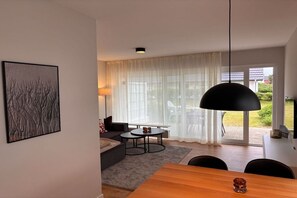 Ein modernes Wohnzimmer mit einer Couch, einem runden Tisch und einem Esstisch. Ein großes Fenster mit durchsichtigen Vorhängen bietet Blick ins Freie.