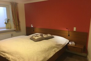 A bedroom with a bed, pillows, bedside table, and a window with curtains. - Landhaus Wattwiesen (Mediterranean) (Rantum)