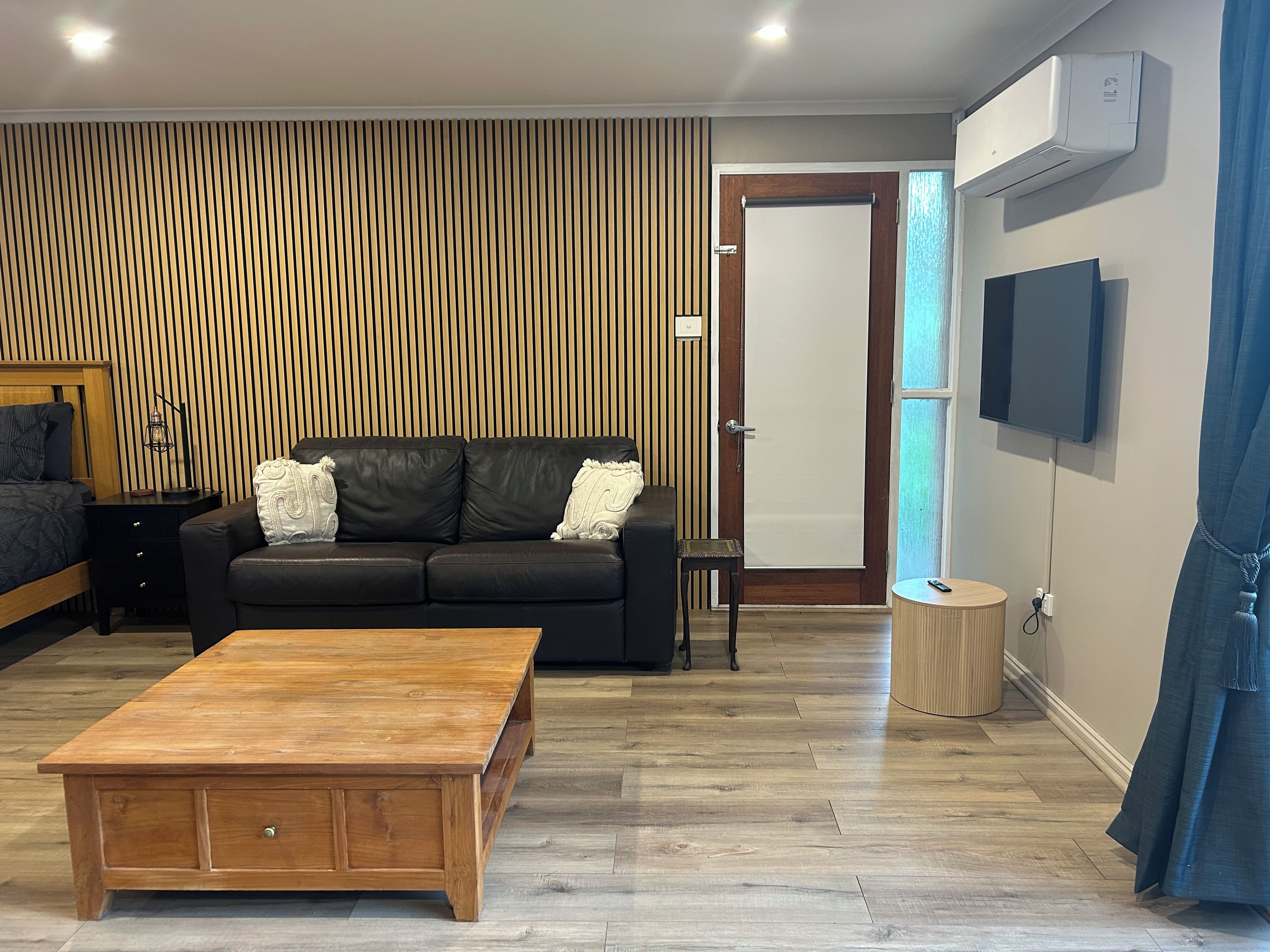 A modern living room with a wooden coffee table, black leather sofa, and a TV mounted on the wall.
