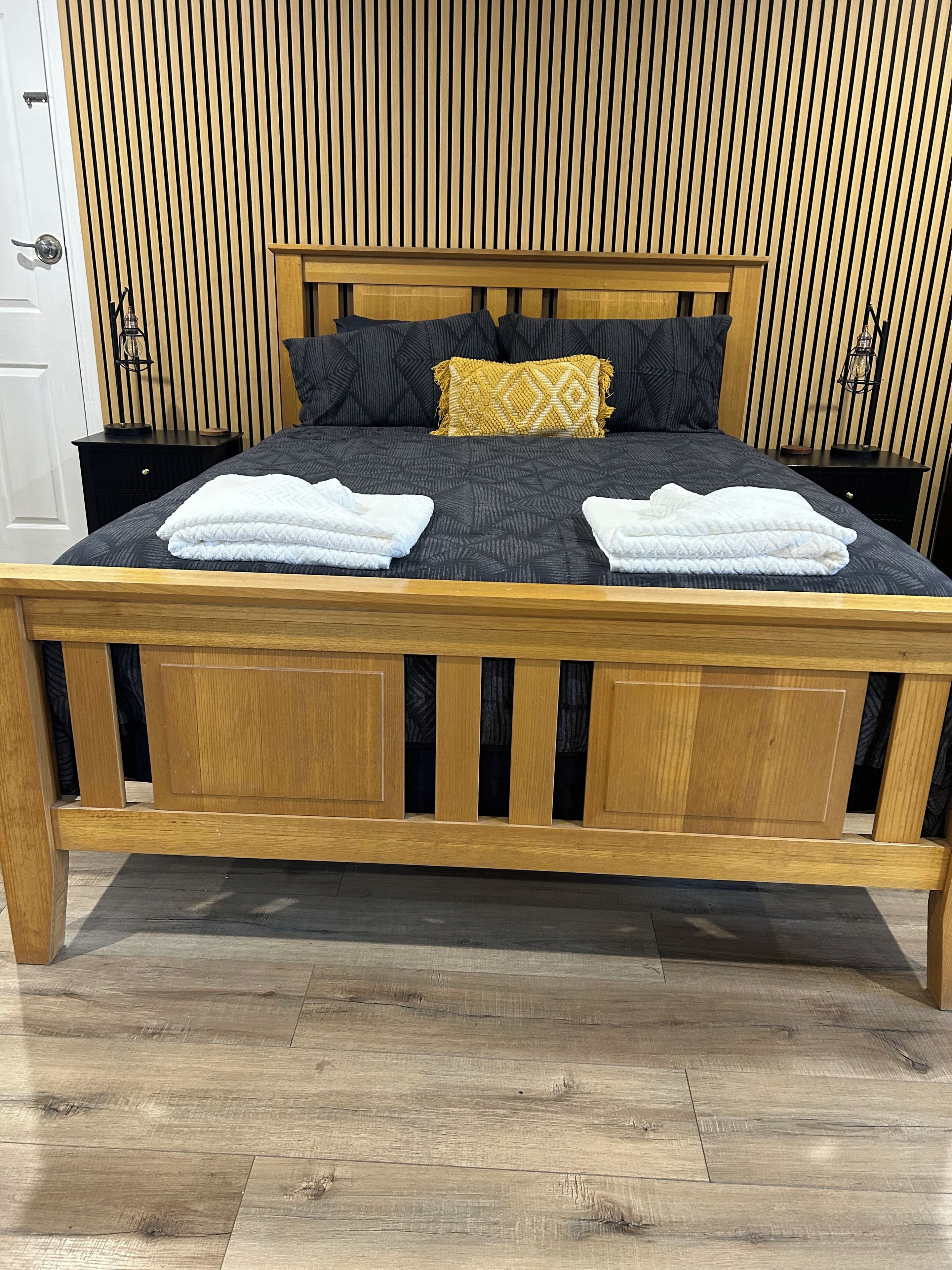 A wooden bed with a dark patterned bedspread, two white towels, and a decorative pillow.