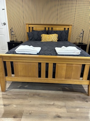Un lit en bois recouvert d’une couverture à motifs sombres, deux serviettes blanches et un coussin décoratif.