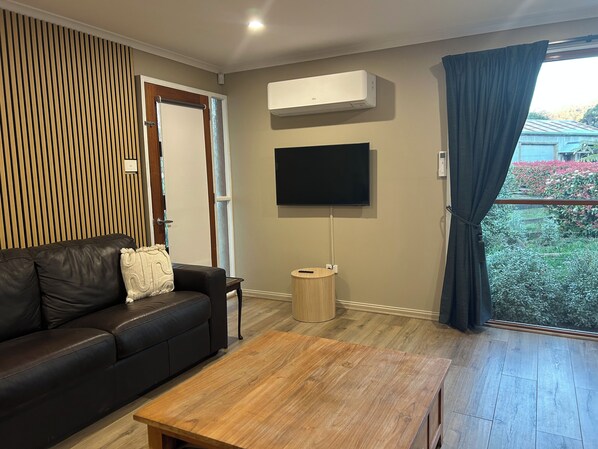 Una sala de estar moderna con un sofá de cuero negro, una mesa de centro de madera, un televisor de pantalla plana montado en la pared y un ventanal con vista a zonas verdes.