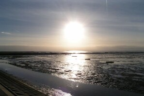 Ein Strand mit tiefstehender Sonne am Horizont, deren Licht sich im Wasser spiegelt.