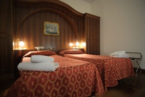 A hotel room with two beds, each with a red patterned bedspread and white towels. There is a wooden headboard with a painting above it and two bedside lamps. - Hotel Grande Bretagne (Montecatini Terme)