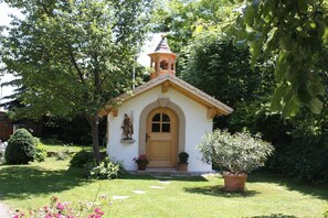 Eine kleine Kapelle mit einem Glockenturm, umgeben von einem gepflegten Garten mit verschiedenen Pflanzen und einer Statue.