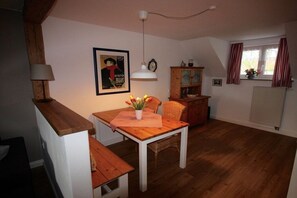 Ein gemütliches Wohnzimmer mit einem Holztisch, einer Bank und einem Esstisch. Eine Hängeleuchte, ein Schrank und ein Fenster mit Vorhängen sind ebenfalls vorhanden.