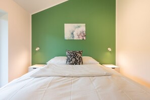 A bedroom with a green wall, a white bed, and a floral pillow. There are two bedside lamps and a painting above the bed. - Ferienhaus mit Eigener Sauna und Grossem Garten (Toenning)