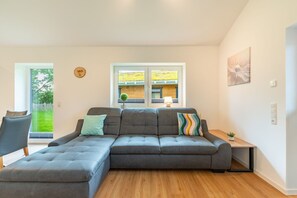 A modern living room with a grey sectional sofa, a small side table, and a window with a view of a garden. - Ferienhaus mit Eigener Sauna und Grossem Garten (Toenning)