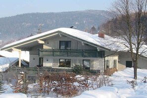 Ein zweistöckiges Gebäude mit Balkon, umgeben von schneebedecktem Boden und Bäumen.