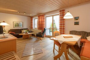 A cozy living room with a sofa, armchair, and table, wooden flooring, and large windows with curtains. - Holiday apartment in Bodenmais with sauna (Bodenmais)