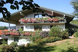 A two-story building with a balcony on the upper floor, surrounded by a garden with various plants and flowers. - Holiday apartment in Bodenmais with sauna (Bodenmais)