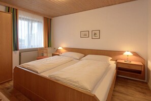 Een tweepersoonsbed met een houten hoofdeinde, twee nachtkastjes met lampen, een raam met gordijnen en een radiator.