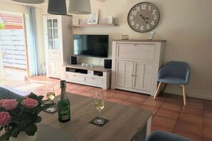 A living room with a flat-screen TV, white cabinets, a clock, a blue chair, and a table with a bottle and glass. - Holiday home with 2 bedrooms (Wangerland-Hooksiel)