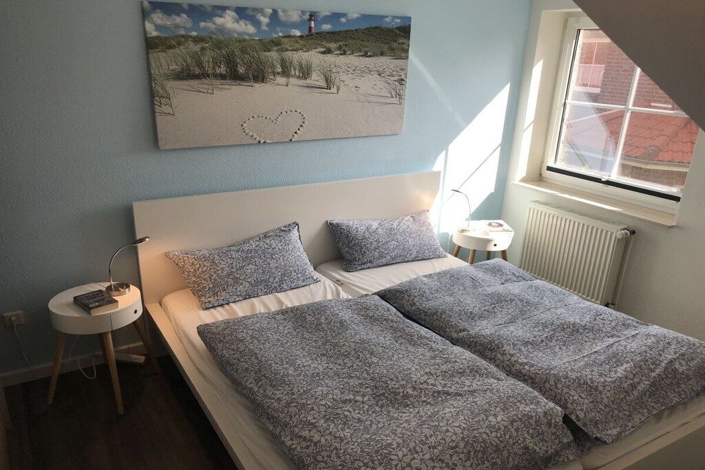A bedroom with a bed, a nightstand, a lamp, a window, and a beach-themed painting on the wall.