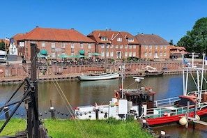 A riverside view with a row of brick buildings, a dock with boats, and a green umbrella. - Holiday home with 2 bedrooms (Wangerland-Hooksiel)