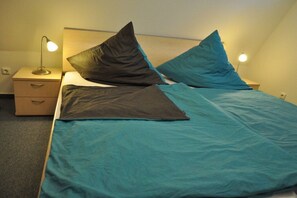 A single bed with teal bedding and two pillows, a bedside table with a lamp, and a drawer unit. - Holiday home Seemuschel (Cuxhaven Duhnen)