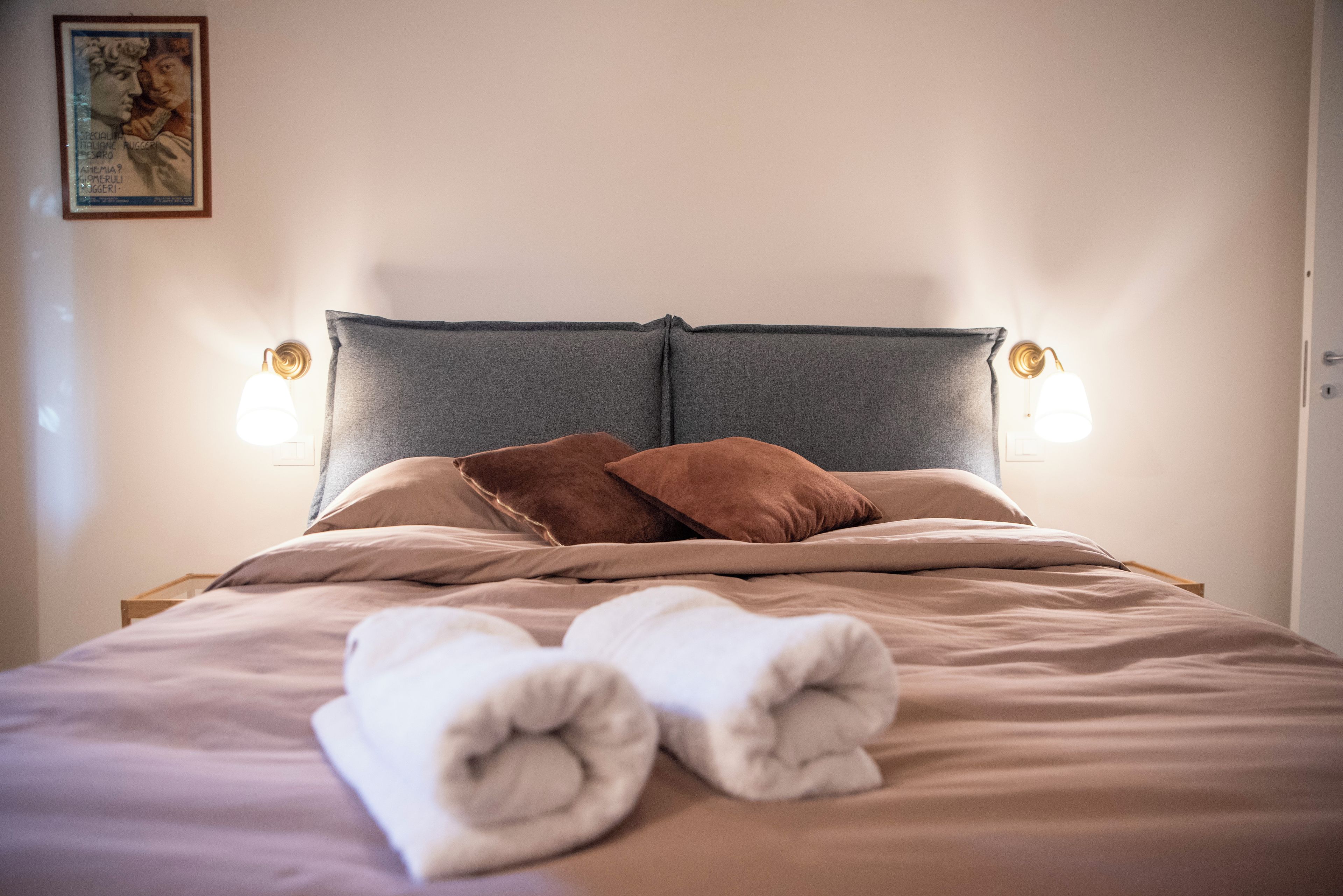 Uma cama bem arrumada com cabeceira cinza, duas luminárias de apoio e um quadro na parede.