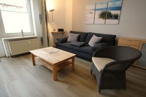 A room with a wooden table, a grey sofa, a wicker chair, a radiator, and a window. - Residence Albatros (Apt. 16) (Cuxhaven-Döse)