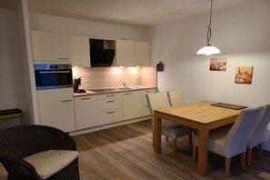 A compact kitchen with white cabinets, a built-in oven, and a dining area with a wooden table and chairs. - Residence Albatros (Apt. 16) (Cuxhaven-Döse)
