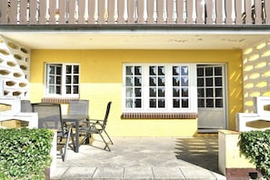 Ein gelbes Gebäude mit zwei Fenstern und einer Tür, eine Terrasse mit Tisch und Stühlen sowie ein weißer Zaun.