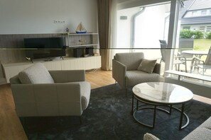 A modern living room with a sofa, armchair, and coffee table, a flat-screen TV, and a view of the outdoors. - House Carola Apartment 15 (Wenningstedt-Braderup)