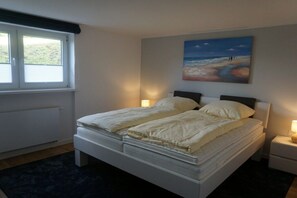 Ein Doppelbett mit weißer Bettwäsche, ein Nachttisch mit Lampe, ein Fenster mit Jalousien und ein Gemälde mit einer Strandansicht an der Wand.