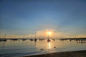 Ein ruhiges Gewässer mit mehreren vor Anker liegenden Booten, einem klaren Himmel und einer sichtbaren Sonne am Horizont.