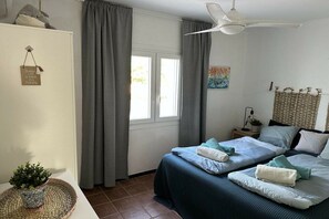 A bedroom with two beds, grey curtains, a ceiling fan, a small table with a plant, and a wall-mounted artwork. - Holiday home with 2 bedrooms (Cala Llombards)