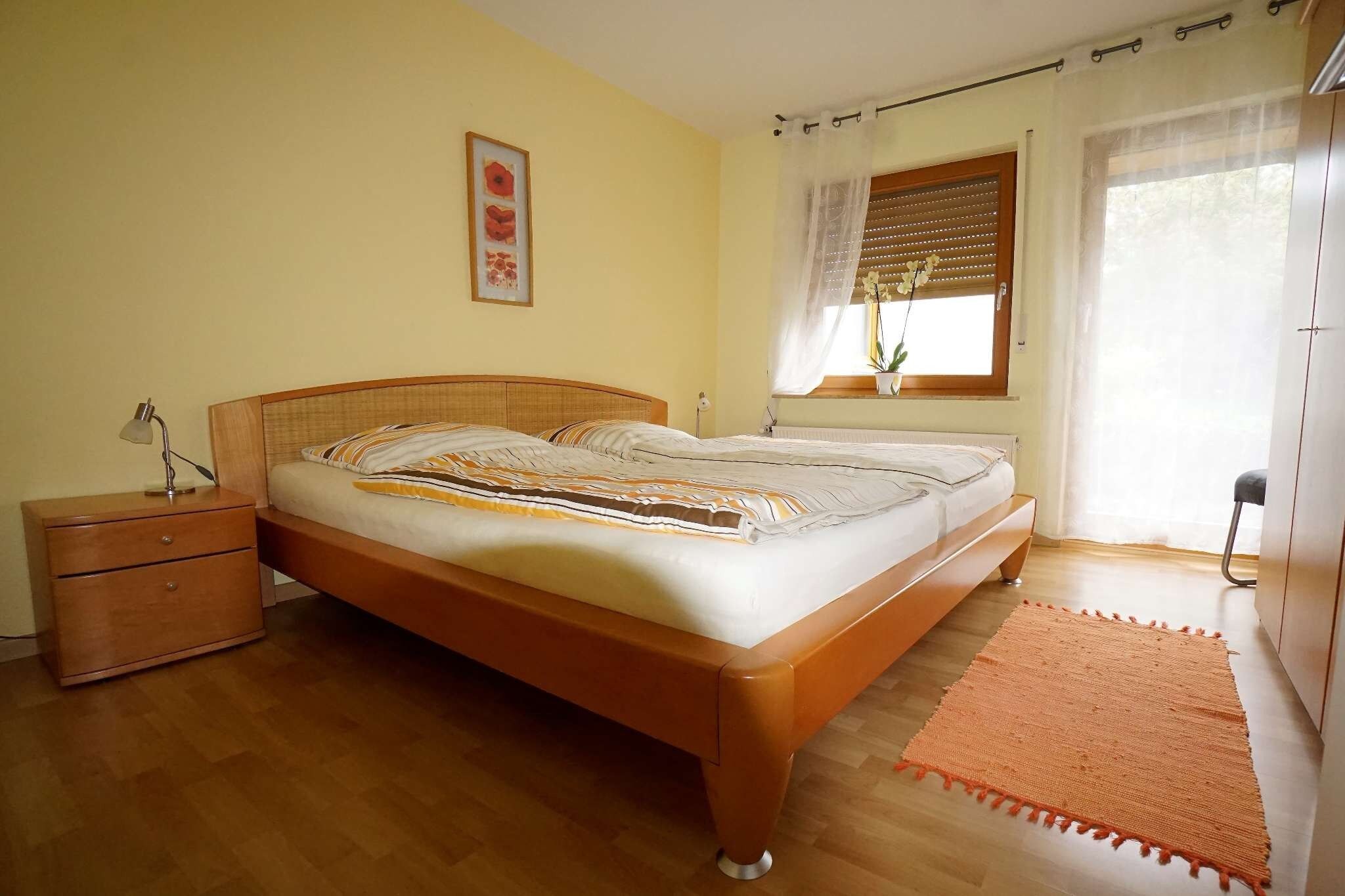 Ein Schlafzimmer mit einem hölzernen Bett, einem Nachttisch, einer Nachttischlampe, einem Fenster mit Jalousien und einer Blumentopfpflanze.
