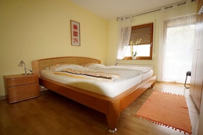 A bedroom with a wooden bed, a nightstand, a bedside lamp, a window with blinds, and a potted plant.