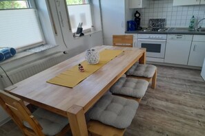 Une cuisine avec des meubles blancs, une table en bois surmontée d’une nappe jaune et une fenêtre avec des stores.