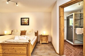 A hotel room with a wooden bed, a bedside table with a lamp, a framed picture on the wall, and a shower area with a glass door. - 4 Country House Farmer (Bodenmais)
