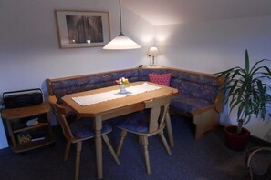 Ein Zimmer mit einem Holztisch, zwei Stühlen, einem blauen Sofa mit einem gemusterten Kissen, einem gerahmten Bild an der Wand, einer Lampe und einer Topfpflanze.