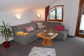 Ein Wohnzimmer mit grauem Sofa, einem Holztisch, einer Topfpflanze und einem Fenster mit Vorhängen.