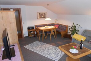 Ein kompakter Wohnraum mit Esstisch, Stühlen, einer Couch, einem Fernseher und einem kleinen Tisch mit Flasche und Blumen.