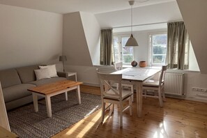 A modern living space with a sofa, a wooden coffee table, a dining table with chairs, a lamp, and a view of the outdoors through a window with curtains. - Holiday apartment with 2 bedrooms (Norddorf)
