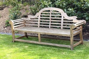 A wooden bench with a backrest and armrests, situated on a grassy area with a hedge in the background. - Holiday apartment with 2 bedrooms (Norddorf)