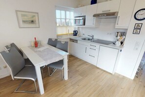 Een kleine keuken met witte kastjes, een eettafel met stoelen, een raam met zonwering en een klok aan de muur.