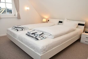 Een tweepersoonsbed met witte lakens, een nachtkastje met een lamp en een raam met gordijnen.