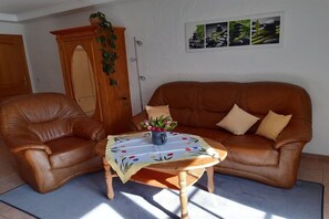 A living room with a brown leather sofa, a wooden coffee table with a floral tablecloth, and a cabinet with greenery. - Holiday apartment with 1 bedroom (Walsdorf-Zilsdorf)