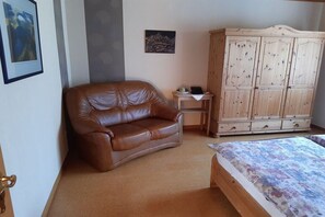 A room with a brown leather sofa, a wooden cabinet, a bed with a floral bedspread, and two framed pictures on the wall. - Holiday apartment with 1 bedroom (Walsdorf-Zilsdorf)